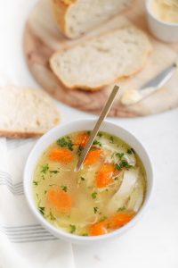 bowl of chicken soup with with bread on the side on wooden cutting board