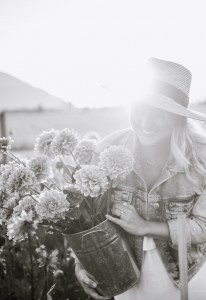 Monika Hibbs black and white smiling woman holding bucket of flowers wearing a denim jean jacket, fresh white dress with sun hat and sun rays in the background