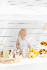 girl behind a lemonade drink stand with 25 cents sign, a basket of lemons and donuts