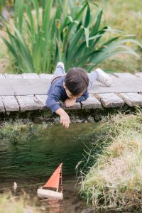 boy on bridge playing with toy boat floating down a stream