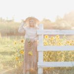 Blonde Woman wearing country girl sweater hat and floral skirt standing in front of sunflowers by a white fence in golden hour
