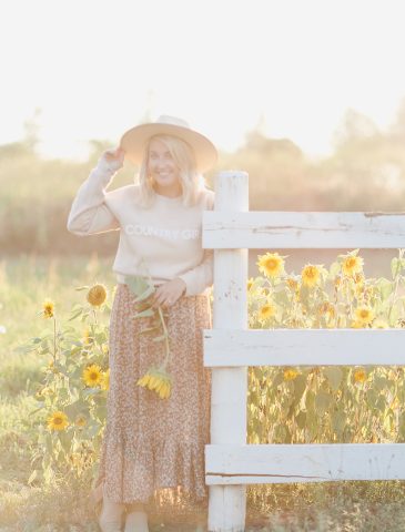 Blonde Woman wearing country girl sweater hat and floral skirt standing in front of sunflowers by a white fence in golden hour