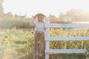 woman wearing a beige hat, country girl sweater and floral print skirt standing in front of sunflower farm